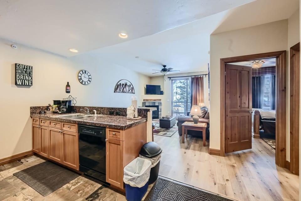 A modern open-concept kitchen and living area with wooden cabinets, a dishwasher, wall decor, and a view of a bedroom. The living room features a sofa, coffee table, and a wall-mounted TV.