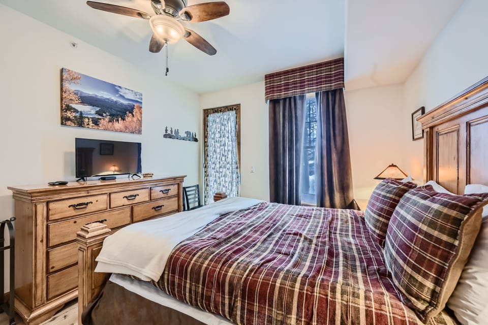 A bedroom with a double bed, plaid bedding, wooden dresser topped with a TV, and a ceiling fan. Decor includes landscape photos and a lamp on the bedside table. A window with curtains is on the right.