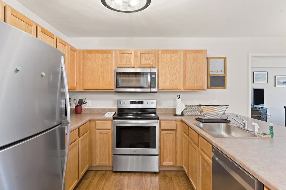 A modern kitchen with light wooden cabinets, stainless steel appliances, and a double sink next to a countertop with a dish rack. The floor is wooden, and there is a doorway to another room.