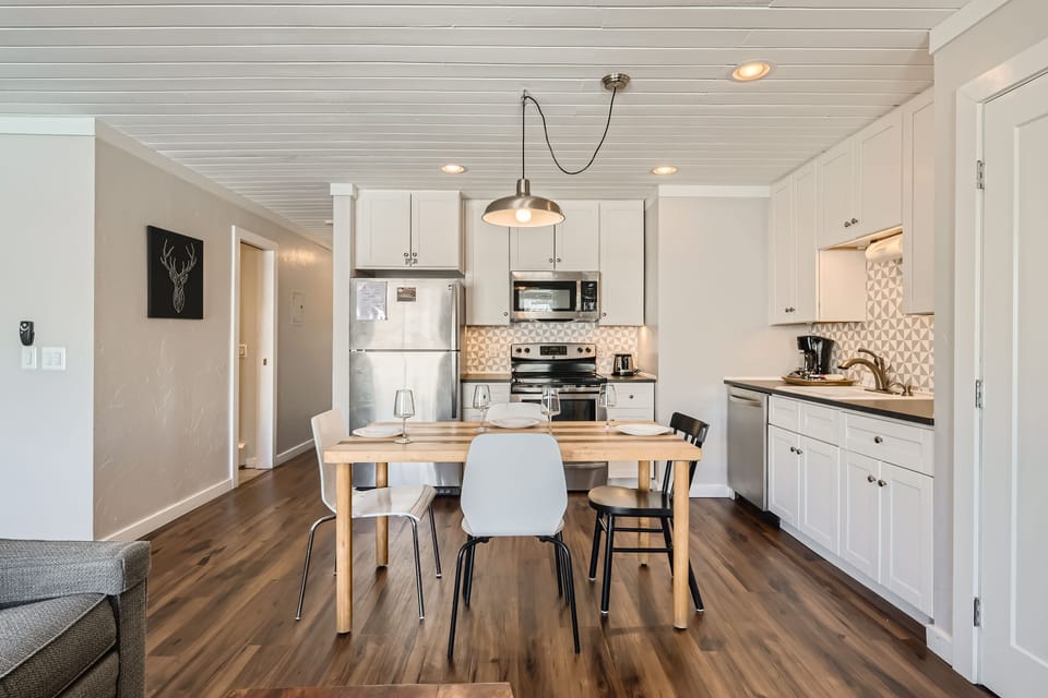 Modern kitchen with white cabinets, stainless steel appliances, wood floors, and a dining table with four chairs set for a meal. Wall art and pendant lighting are visible.