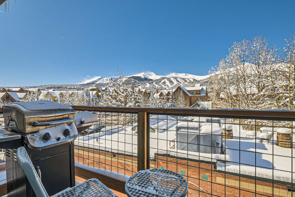 Outdoor balcony with a grill and metal table set, overlooking a snowy mountain landscape and ski resort buildings under a clear blue sky.