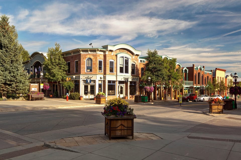 A quaint downtown corner features historic buildings, shops, and a central street, with planters filled with flowers. A few people, trees, and parked cars enhance the scene under a partly cloudy sky.