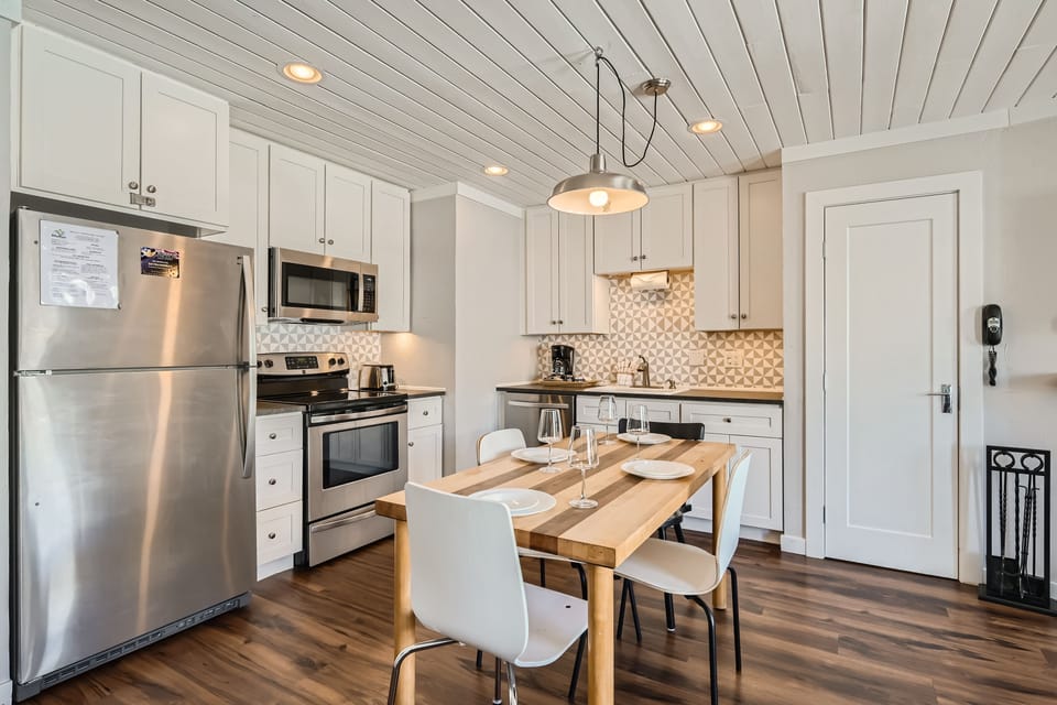 A modern kitchen featuring stainless steel appliances, wood flooring, a central wooden dining table set for two, and white cabinets with patterned backsplash under recessed lighting.