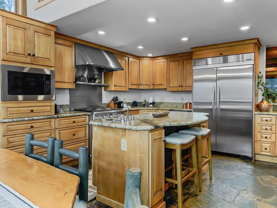 A modern kitchen features wooden cabinets, a stainless steel refrigerator, a built-in microwave, an island with bar stools, and a stone tile floor.