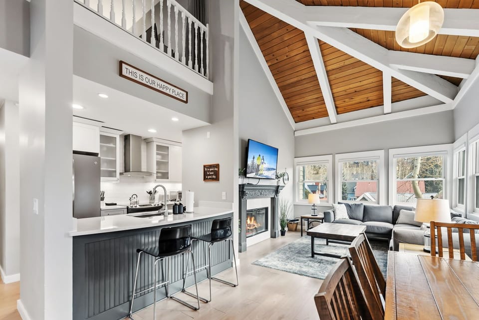 The open-space living/dining/kitchen is a stunning area of the condo filled with charming details such as exposed wood and white beams, a beautiful fireplace mantle, and a wall of windows.