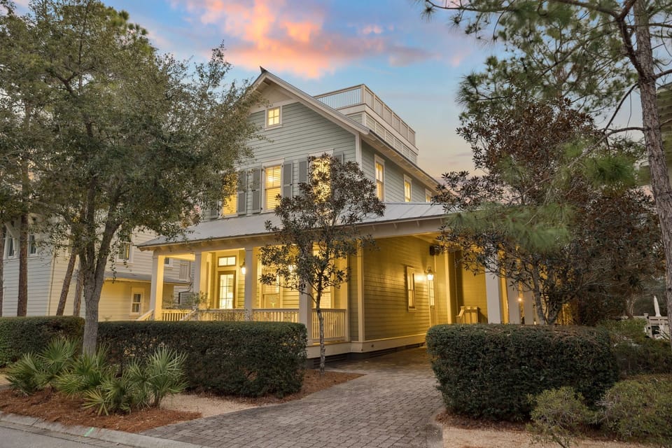 A beautiful coastal home welcomes guests with warm lighting and mature landscaping as evening approaches.
