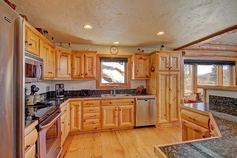 A spacious, rustic kitchen featuring wooden cabinetry, stainless steel appliances, granite countertops, and a window with a view of snowy mountains and trees.