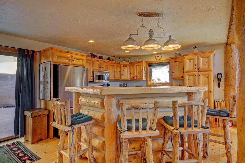 A rustic kitchen features wooden cabinets, a log bar with four high stools, stainless steel appliances, and a quadruple pendant light hanging from the ceiling.