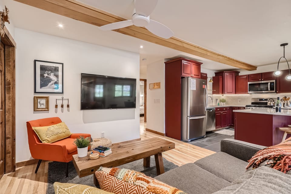 A cozy living area with an orange armchair, a wall-mounted TV, and a wooden coffee table. The kitchen in the background features red cabinets, stainless steel appliances, and pendant lighting.