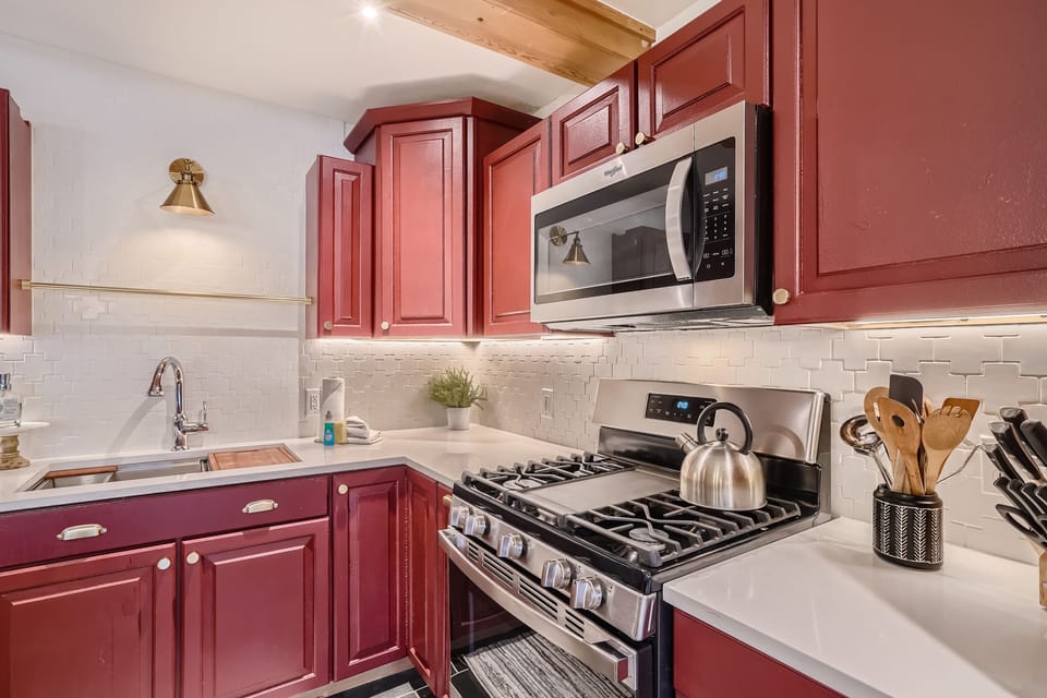 A modern kitchen with red cabinets, stainless steel appliances including a microwave and oven, a kettle on the stove, and a sink under a wall-mounted light. White backsplash tiles and countertops.