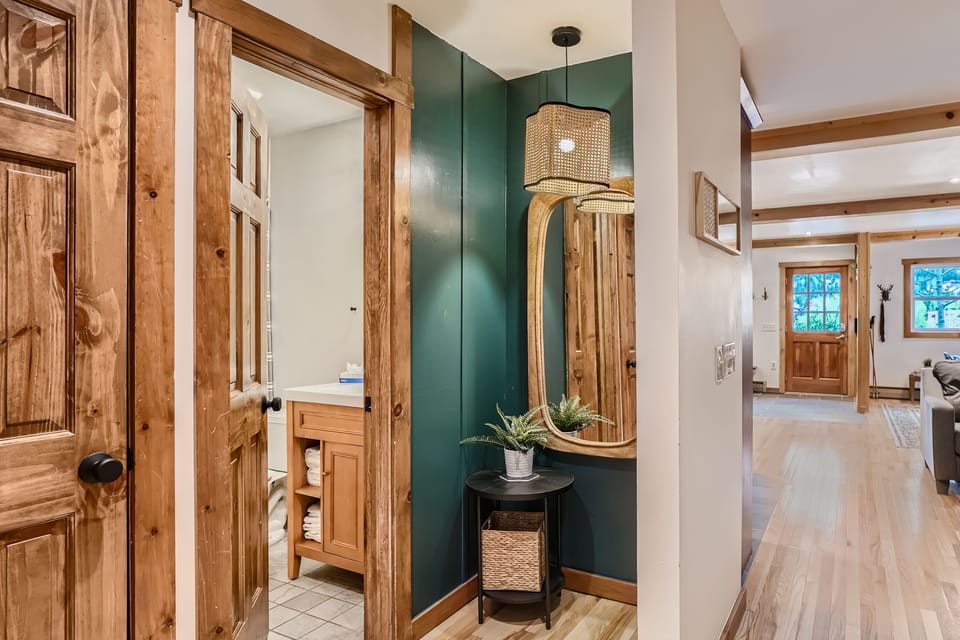 A hallway with wooden trim features a wall-mounted mirror and a small black table with a plant. An open door reveals a bathroom with a wooden vanity. A living area is visible in the background.