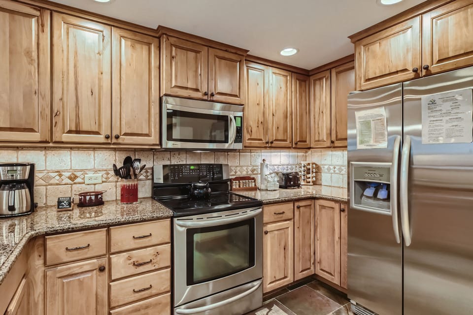 A kitchen with wooden cabinets, stainless steel appliances, a tiled backsplash, and granite countertops. It includes a refrigerator, microwave, stove, coffee maker, and various kitchen tools.