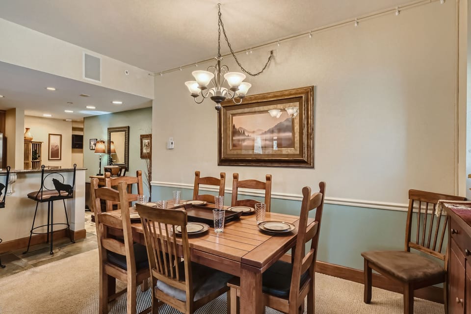 A dining room with a wooden table set for six, adorned with plates and glasses. A chandelier hangs overhead, and a framed landscape picture is on the wall. The background shows a kitchen area.