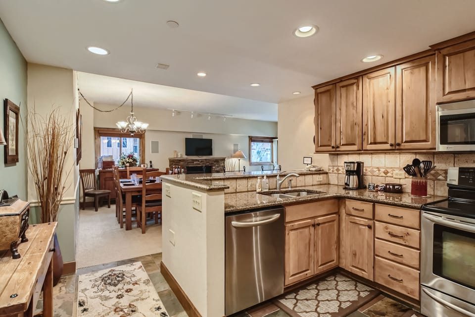 A modern kitchen with stainless steel appliances and wooden cabinets opens into a dining and living area. The space is well-lit with recessed lighting and a chandelier above the dining table.