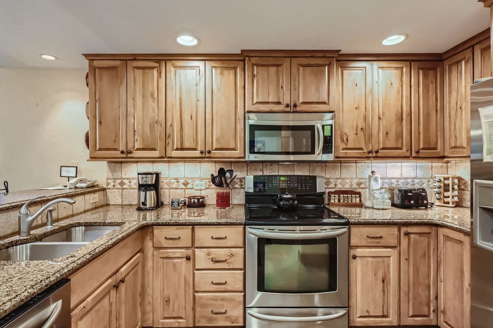 A modern kitchen featuring wooden cabinets, granite countertops, and stainless steel appliances, including a microwave, oven, and double sink. Countertop items include a coffee maker and toaster.