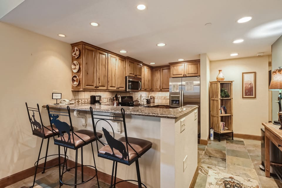 A modern kitchen with wooden cabinets, a granite countertop, recessed lighting, and bar stools. The floor is tiled, and there are decorative items and artworks displayed on the walls and shelves.