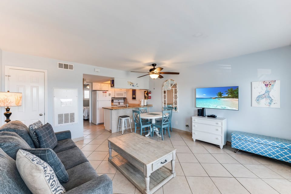 Relax in your bright, coastal-themed living space with comfortable seating, modern kitchen, and tropical beach views on the large TV.