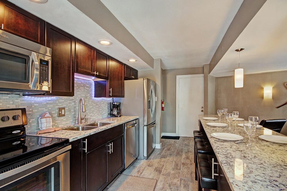 A modern kitchen with stainless steel appliances, dark wood cabinets, granite countertops, and a kitchen island with seating and place settings. A pendant light hangs above the dining area.
