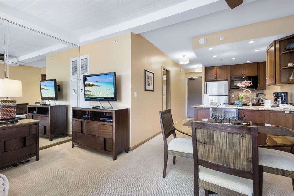 Relax in your open-concept living area with a fully-equipped kitchen, cozy dining for four, and entertainment essentials including a flatscreen TV—all bathed in warm, welcoming natural light.