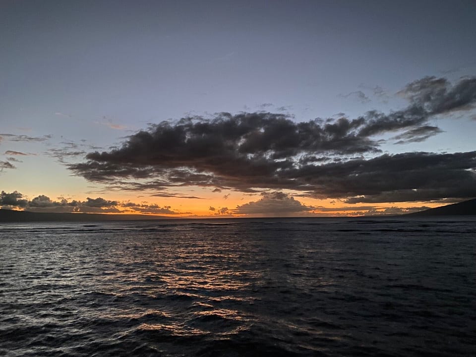 Dramatic sunset paints the sky with golden and amber hues over tranquil waters, framed by distant hills and striking cloud formations along the horizon.