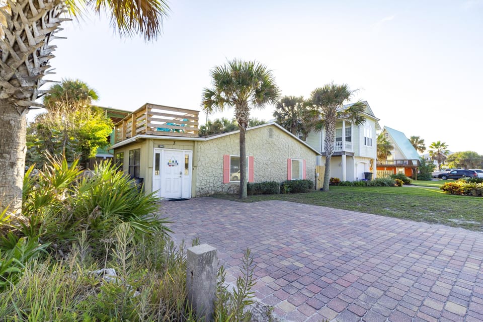 Front exterior view of the house with textured beige walls, coral shutters, and a rooftop deck surrounded by palm trees.