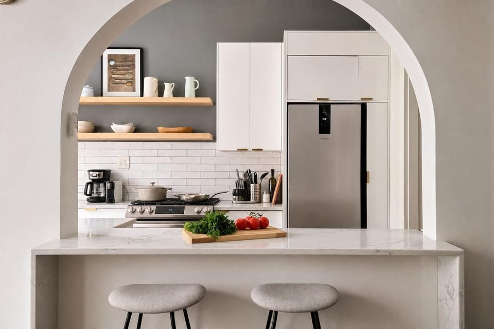 Marble breakfast bar with two gray stools frames a kitchen featuring stainless steel appliances and open wood shelves.