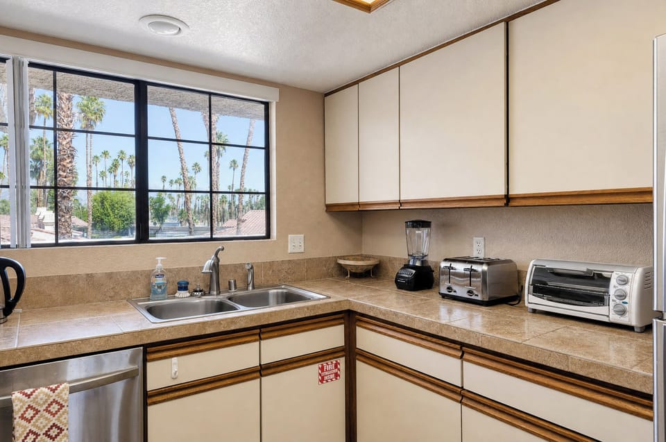 Your fully equipped kitchen features modern appliances and ample counter space for preparing meals, with tropical palm views through large windows that fill the space with natural light.