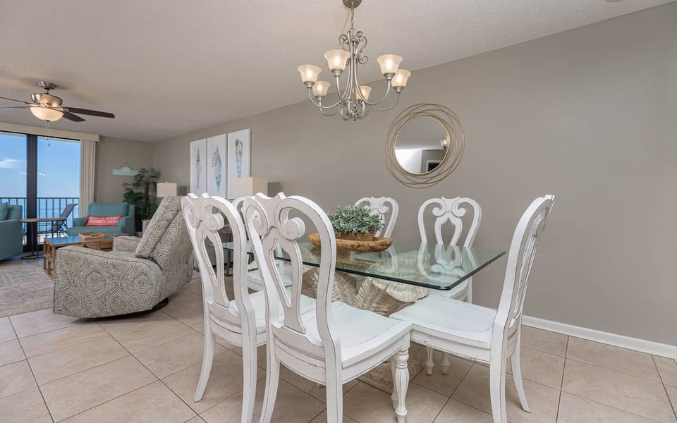 Dining area features a glass table with white chairs, complemented by coastal decor and a view of the balcony