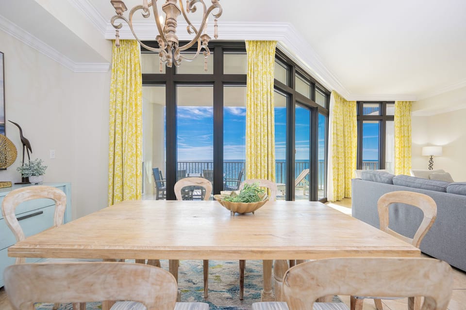 Bright dining area with a wooden table, yellow curtains, and a stunning view of the balcony and serene waters beyond