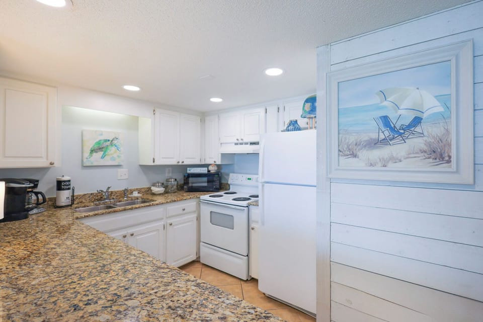 This kitchen features white cabinetry, granite countertops, and a beach-themed artwork, perfect for culinary adventures