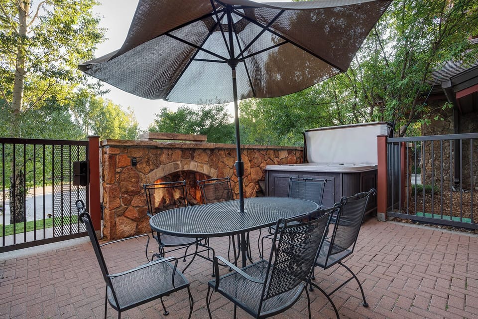 Your private patio sanctuary features elegant dining under a shaded umbrella, with a cozy stone fireplace and relaxing hot tub nearby.