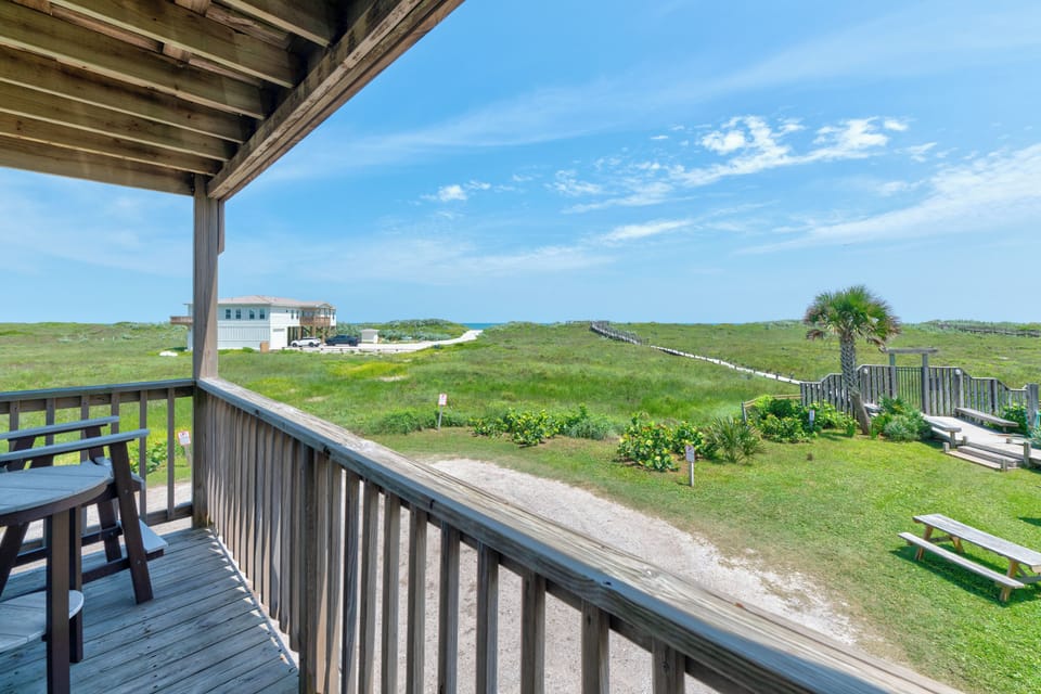A covered wooden balcony overlooks coastal dunes and grassy vegetation with a glimpse of the ocean beyond, creating a peaceful seaside setting with nearby beach access paths and picnic areas.