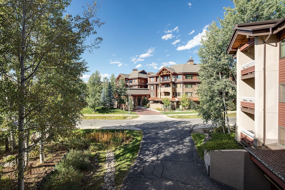 Mountain resort buildings nestled among tall aspen trees under clear blue skies, with paved paths winding through the peaceful property grounds.