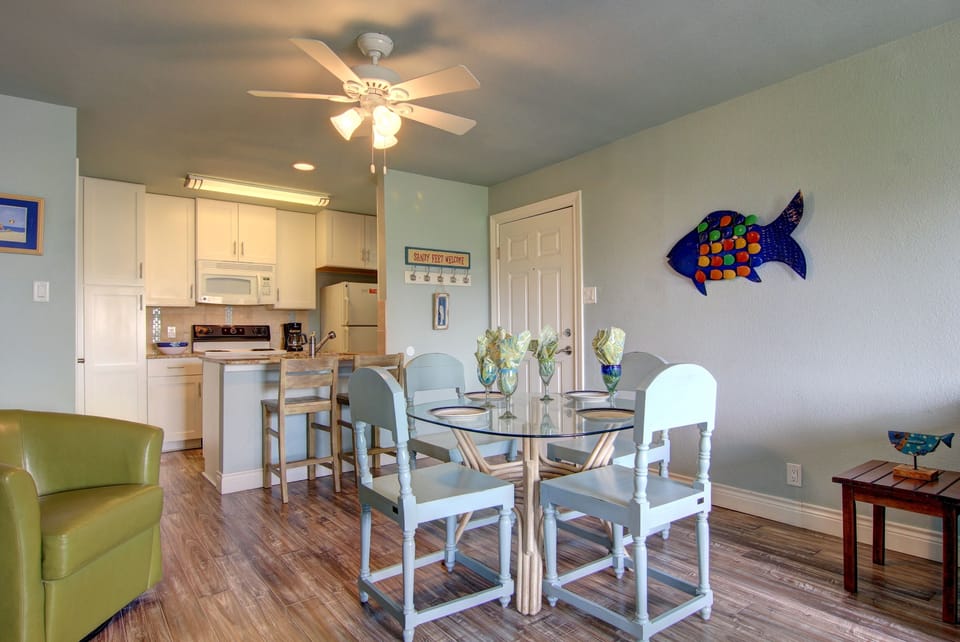 Gather around your glass dining table where coastal charm meets modern comfort. The open kitchen invites easy meal prep while whimsical fish art adds seaside spirit.