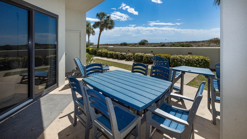 Outdoor patio area features a large blue table surrounded by matching chairs, with views of lush greenery and the beach