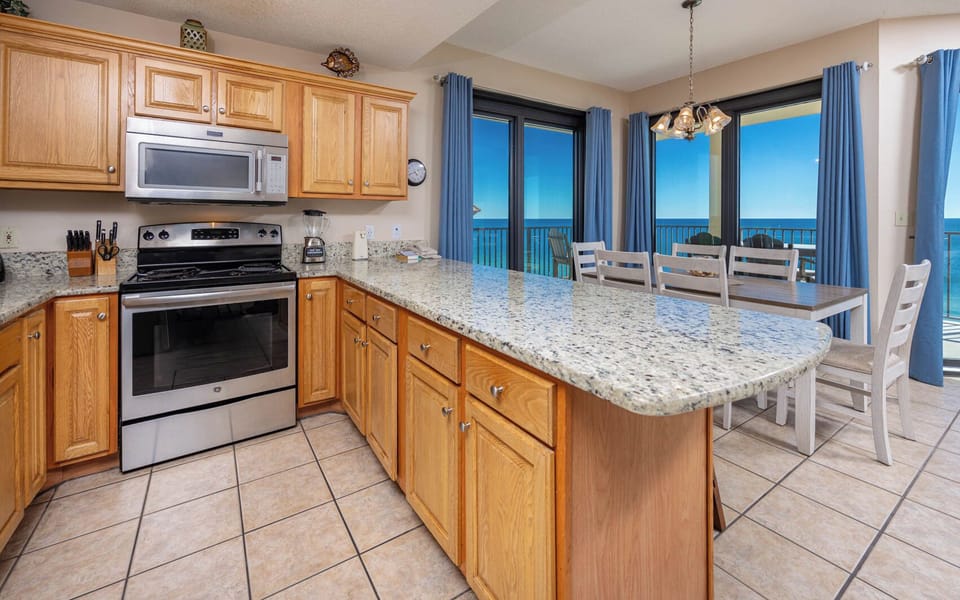 The modern kitchen features warm wood cabinetry, a spacious granite countertop, and a dining area with beach front views