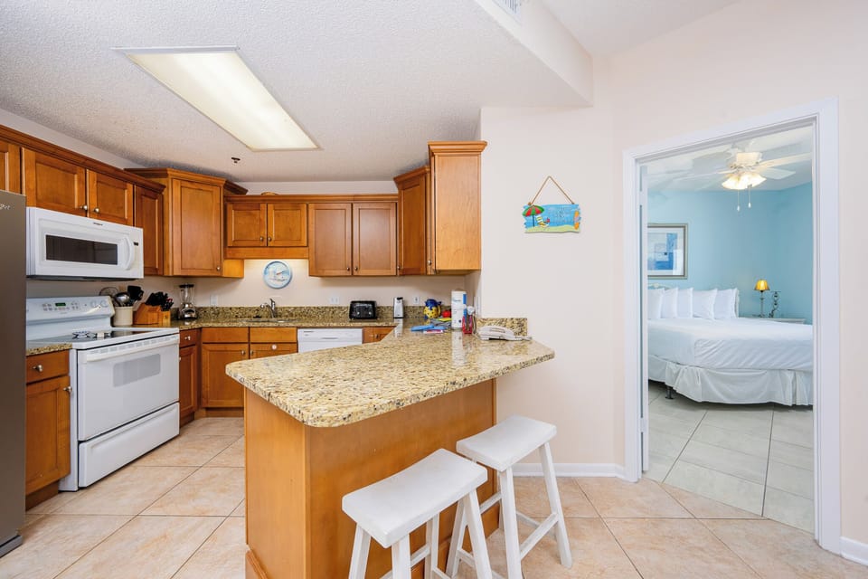 The kitchen features warm wood cabinetry, a granite breakfast bar with seating, and a view into the master suite