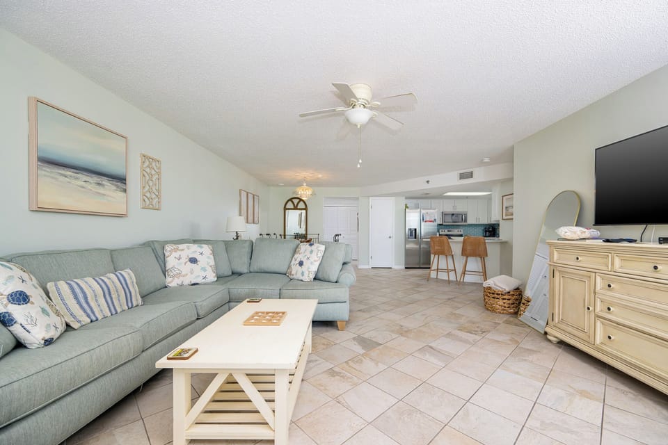 The airy living room features a light blue sectional sofa with decorative pillows, a white coffee table, and a flat-screen TV