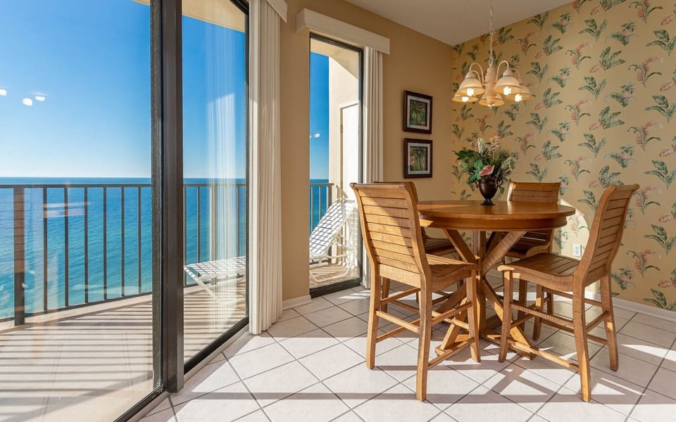 Bright dining area with a round wooden table, four chairs, and a stunning view of the beach through large sliding glass doors