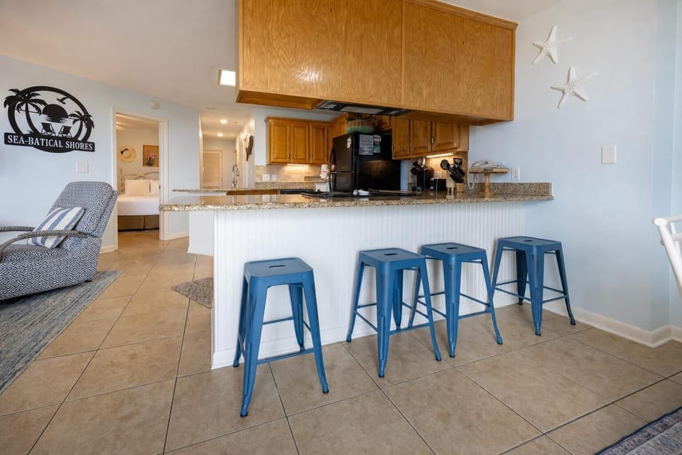 A cozy breakfast bar with blue stools complements the kitchen's wooden cabinetry, leading to a welcoming bedroom