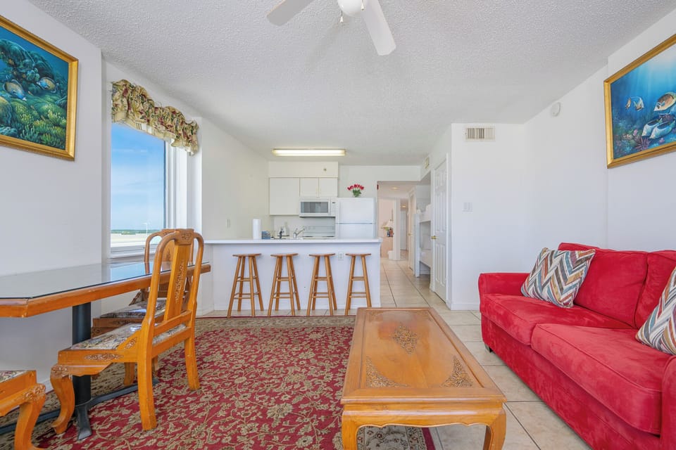 The living area features a cozy red sofa, a TV, and a dining table with a beautiful beachfront view