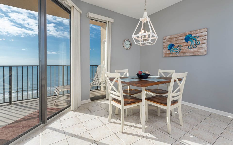Bright dining area features a wooden table with white chairs and a view of the beach through large sliding glass doors