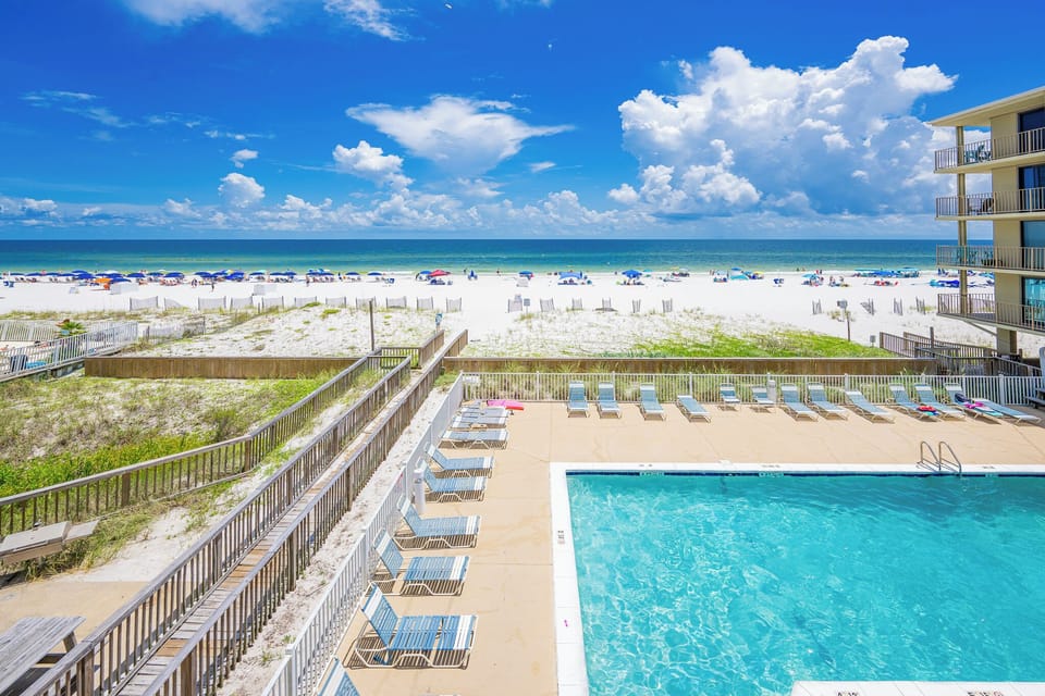The view showcases a sparkling pool surrounded by lounge chairs, leading to a beautiful sandy beach and vibrant umbrellas