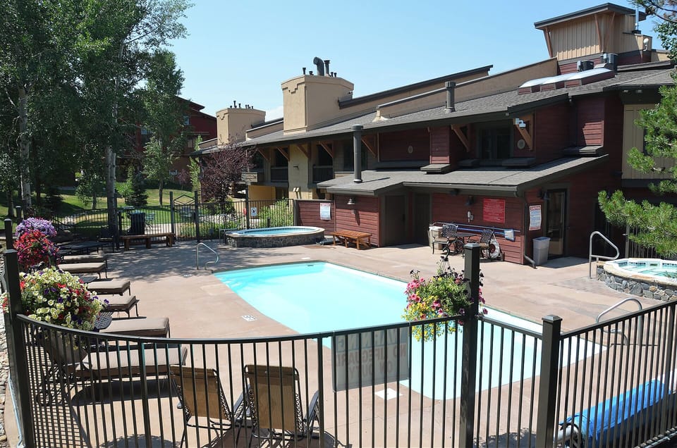 Modern mountain resort complex featuring outdoor swimming pool, hot tub, and contemporary condominium buildings surrounded by mature trees.