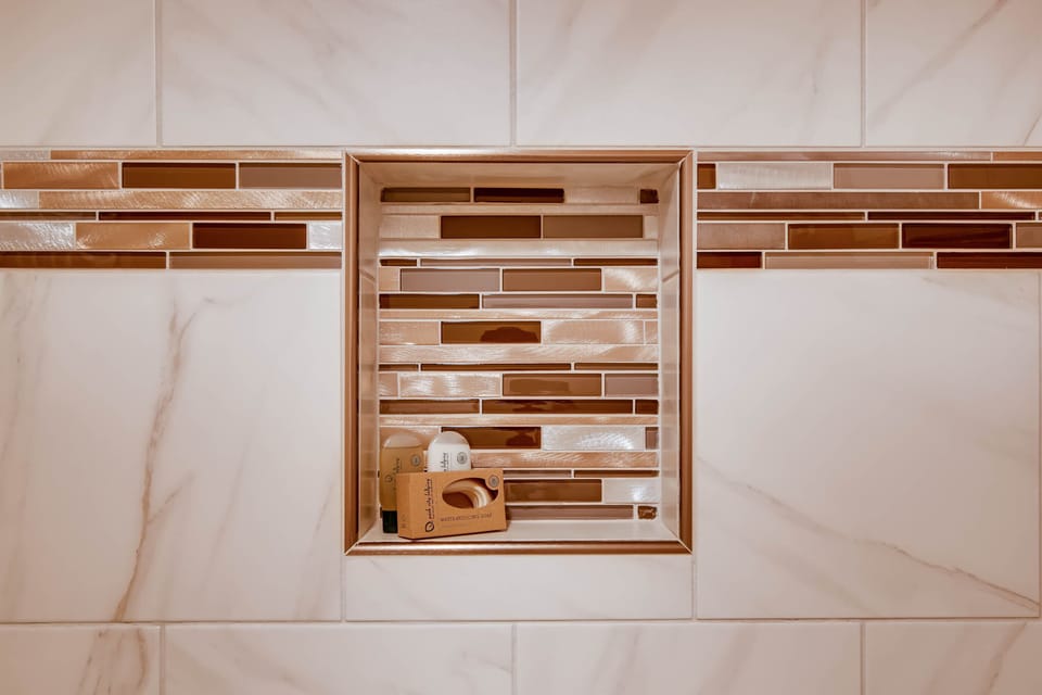 The en suite bathroom has gorgeous white tile, with gold and brown decorative tile lining a shelf in the bathtub surround.
