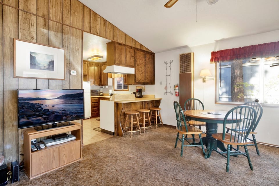 This versatile space features comfortable dining for four, breakfast bar seating, and a cozy lounge area with a flat-screen TV. The wood-paneled walls and open layout create a classic, inviting cabin atmosphere.