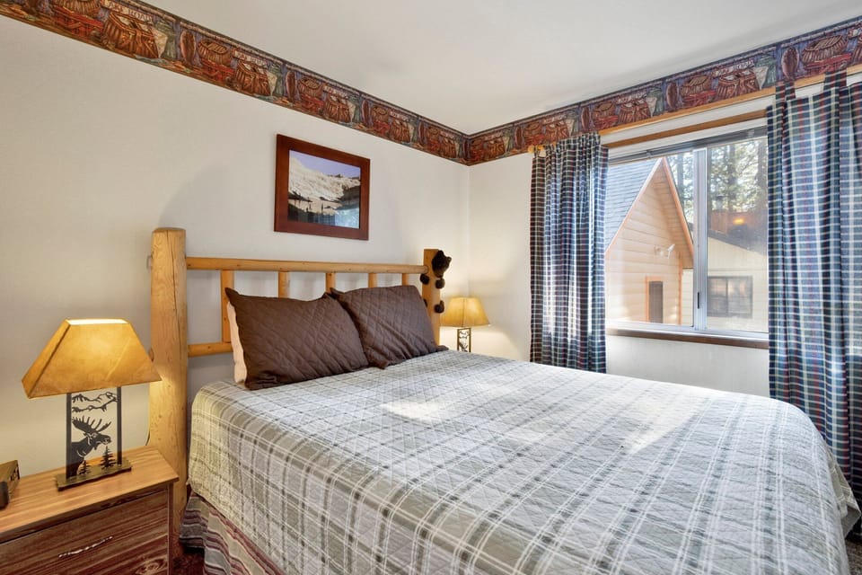 A restful space featuring a queen bed with rustic log framing and decorative mountain-themed borders. Large windows provide plenty of natural light and peaceful views of the surrounding neighborhood.