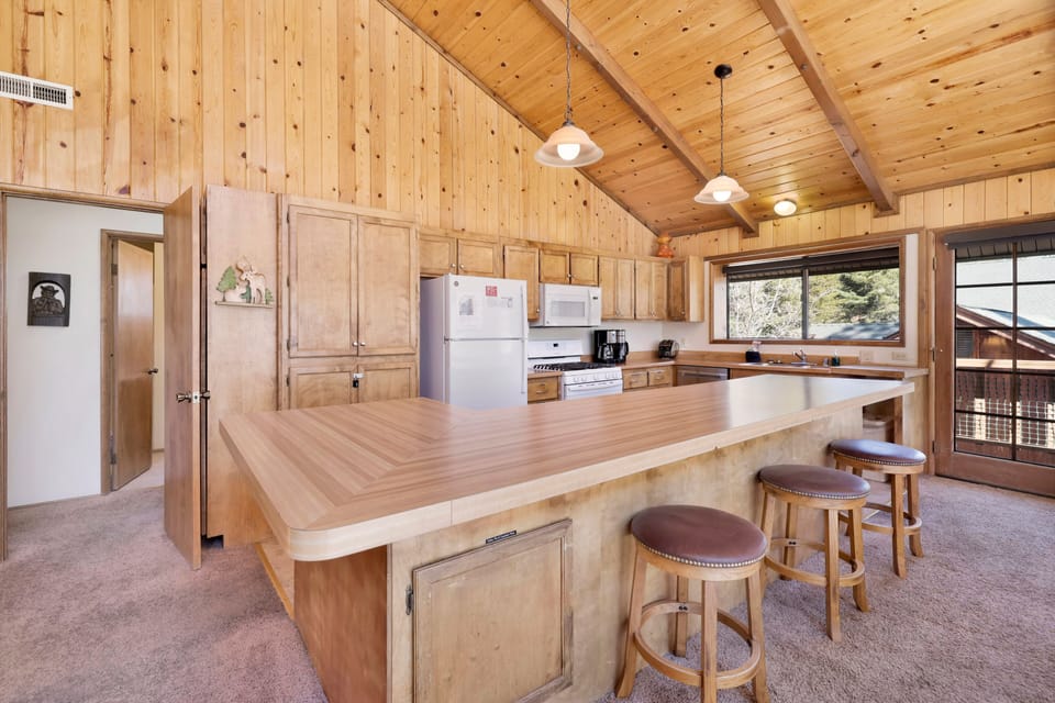 Bright cabin kitchen and dining area with wood ceilings, large island seating, and forest views—ideal for family meals, group cooking, and comfortable mountain getaways.