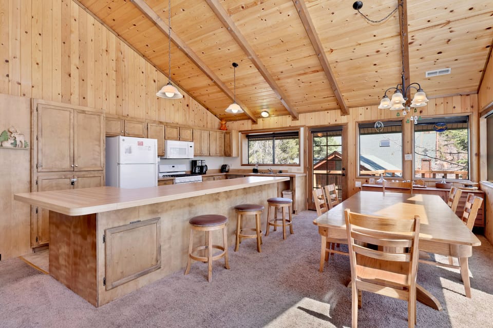 Bright cabin kitchen and dining area with wood ceilings, large island seating, and forest views—ideal for family meals, group cooking, and comfortable mountain getaways.