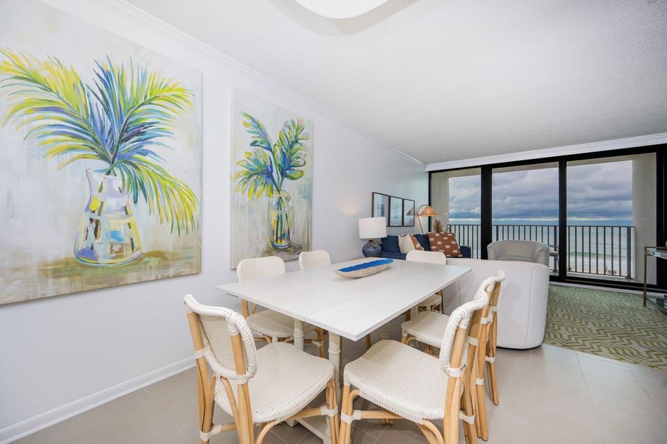 The bright dining area features a stylish white table, comfortable seating, and a stunning beach view through large windows